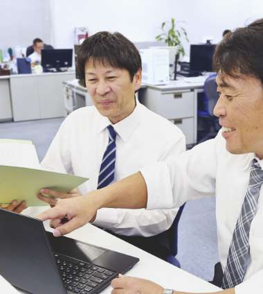2名の男性社員が、ノートPCとファイルを見て楽しそうに何かを相談している写真